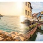 Panoramic split-scene showing Cannes palm-lined promenade and yachts alongside Colmar's colorful half-timbered canal houses at golden hour