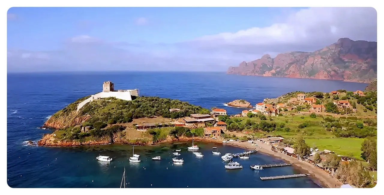 Aerial panorama of a Corsican headland with a small fortress, moored boats and a village backed by hills