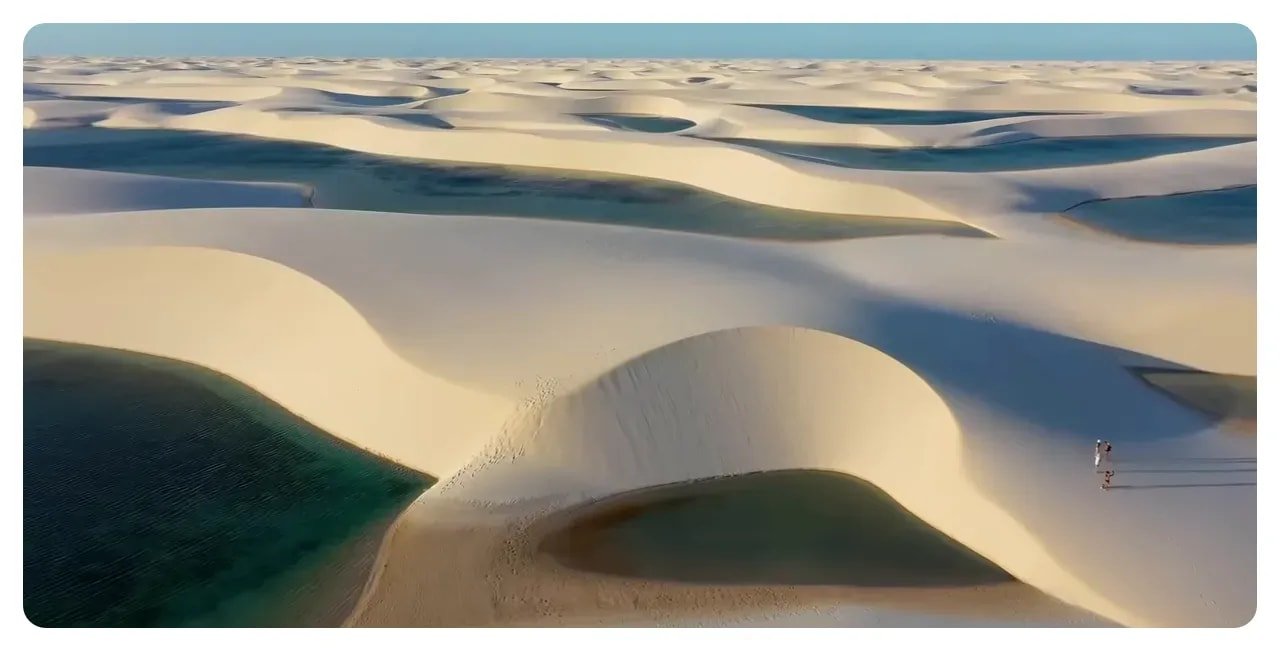Aerial sweep of Lençóis Maranhenses showing rolling white sand dunes, turquoise lagoons and a few visitors on the dune for scale