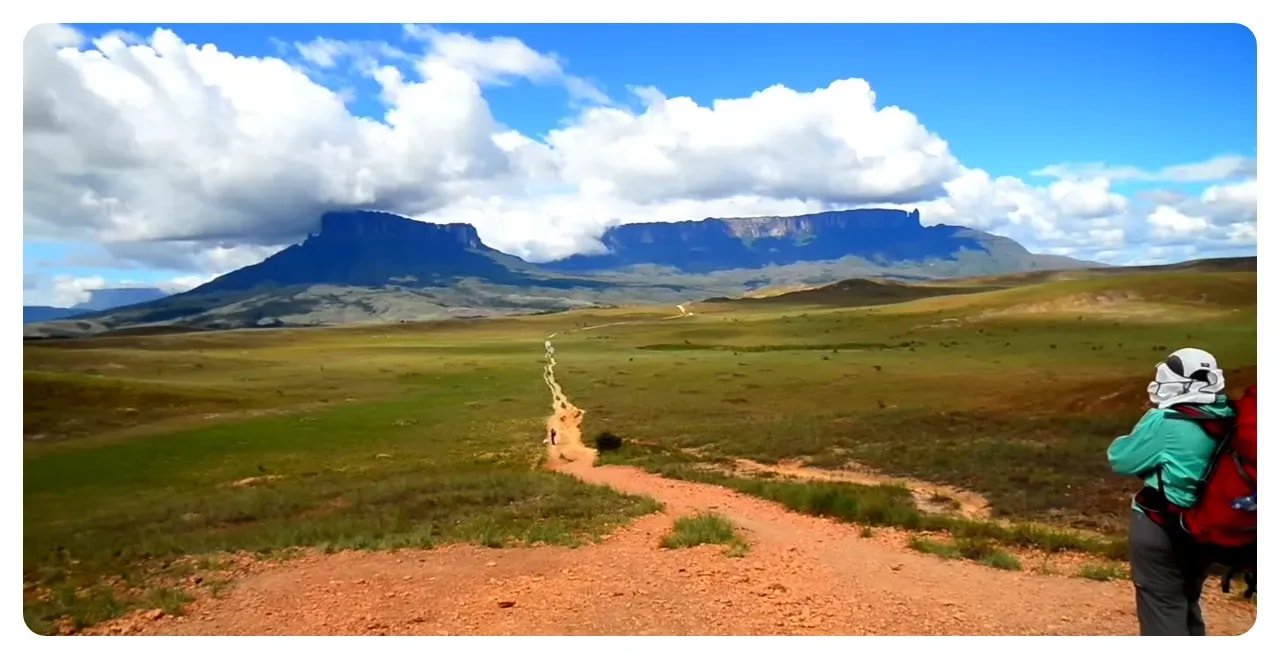 Wide landscape with a dirt trail stretching toward flat‑topped tepui mountains under a dramatic sky; a hiker with a red backpack stands at right.