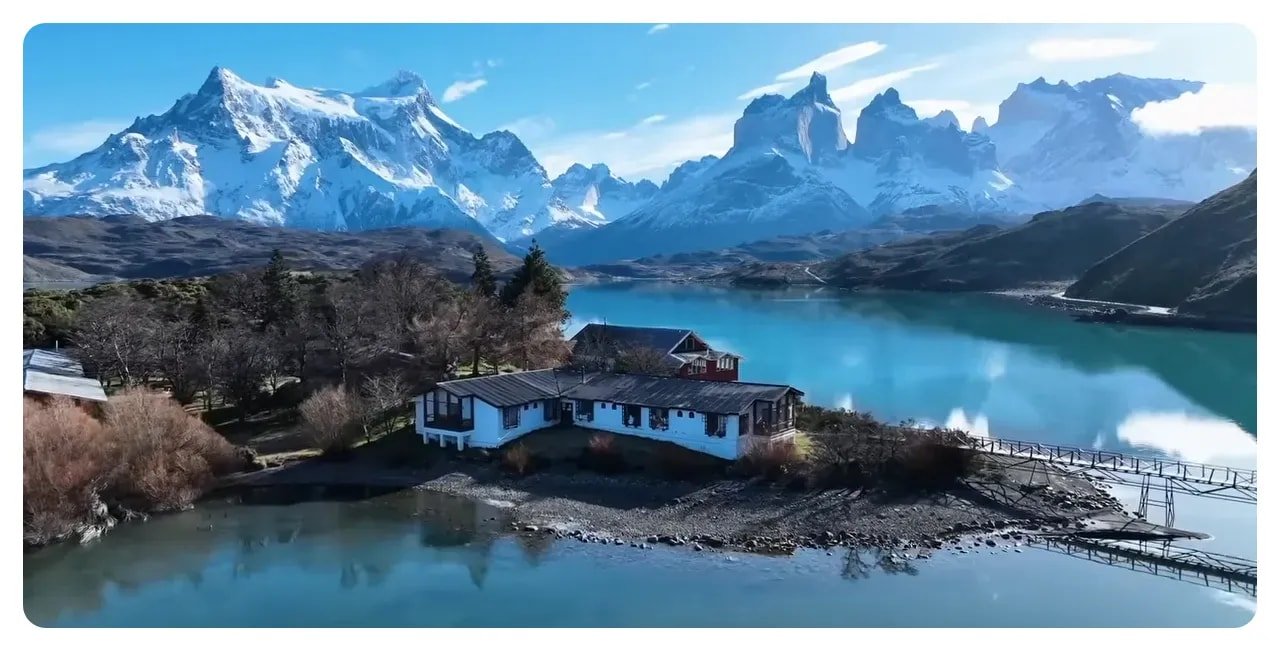 Aerial view of jagged, snow-capped granite peaks reflected in a turquoise lake with a small lodge on the shore.