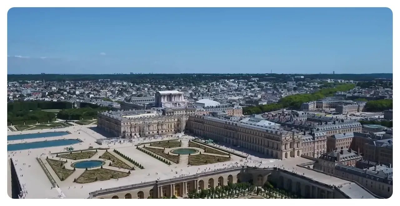 Wide aerial view of the Palace of Versailles showing the palace roofline, parterre gardens and long terraces